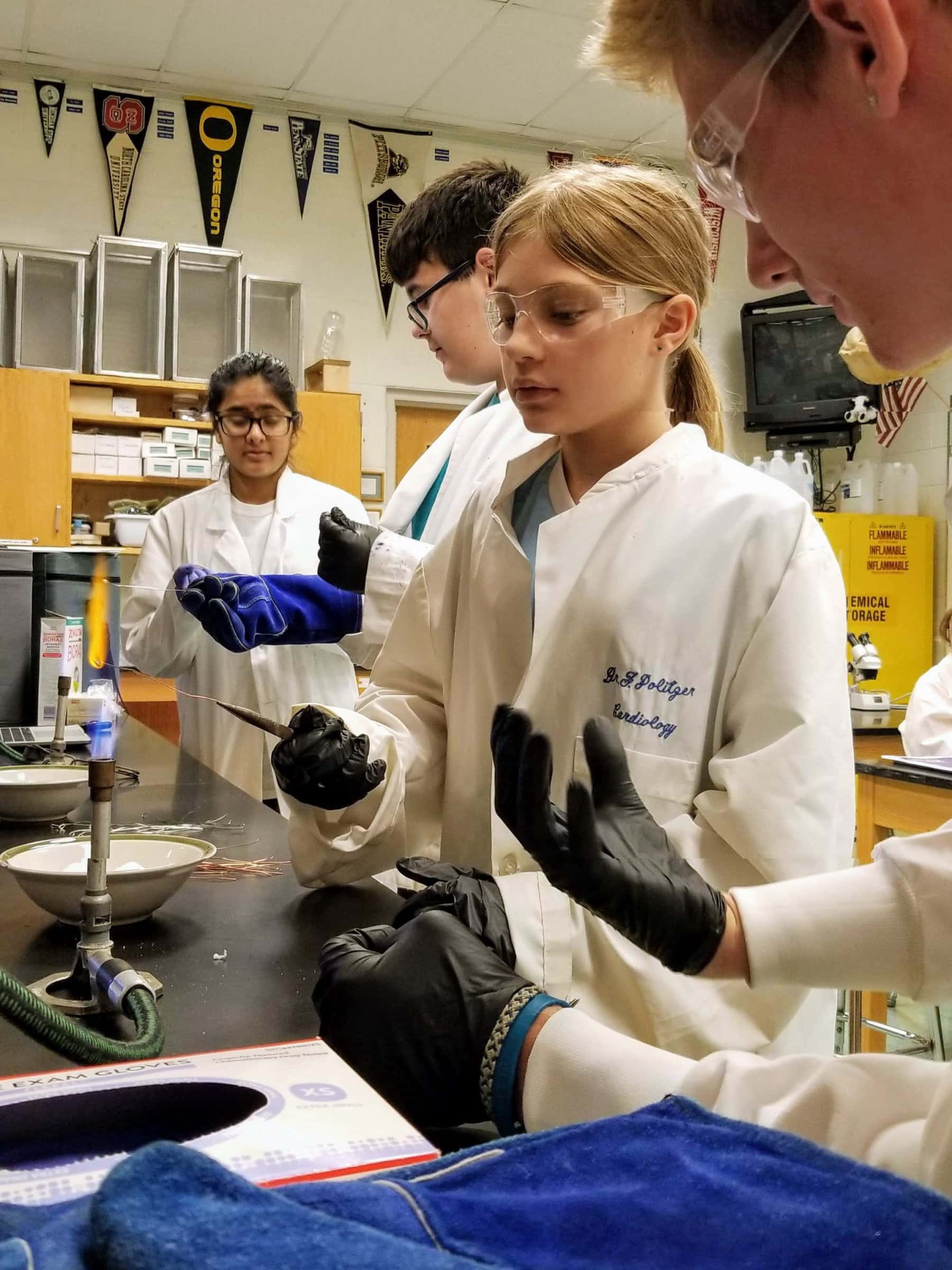 Young blonde girl in lab coat and gloves holds thin wire over a Bunsen burner flame